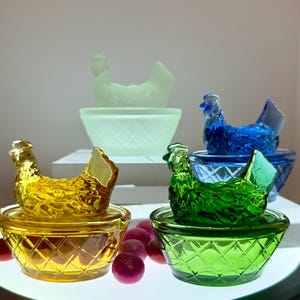 May include: A collection of colorful glass hen-shaped trinket boxes. The hens are in shades of yellow, green, blue, and a pale green, each sitting atop a matching diamond-patterned bowl. The background features a white display.