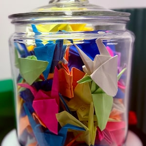 May include: A glass jar filled with colorful origami paper cranes. The cranes are folded in various colors, including red, orange, yellow, green, blue, and purple.