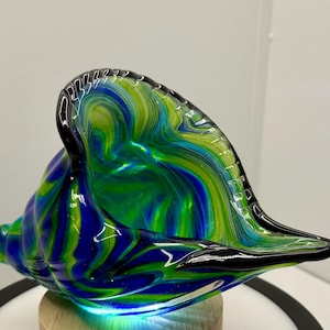 May include: A handcrafted glass seashell sculpture in shades of blue, green, and yellow. The swirled colors create a marbled effect. The shell is resting on a wooden base. The edges are outlined in black.