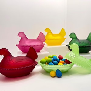 May include: A collection of colorful glass hen-shaped containers, each in a different hue: red, pink, yellow, green, and light green. A bowl of colorful eggs sits in front of the hens. The containers are likely for decorative or storage purposes.