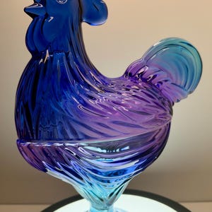 May include: A rooster-shaped glass figurine featuring a vibrant gradient of colors, transitioning from a deep cobalt blue at the head and neck to a rich purple in the center, and fading into a light turquoise blue at the base and tail.