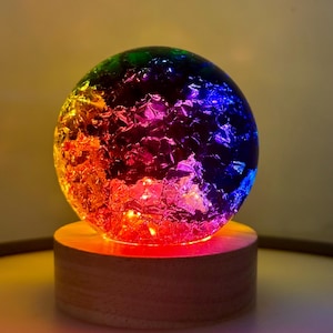 May include: A colorful, illuminated sphere with a textured surface, resting on a round wooden base. The sphere transitions through a spectrum of colors, including red, orange, yellow, green, and blue. The base is approximately 3 inches in diameter.