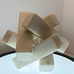 May include: A cream-colored, abstract sculpture composed of interlocking rectangular blocks. The blocks are arranged in a complex, interwoven pattern, creating a visually intriguing and modern decorative piece. The sculpture is made of a smooth, possibly stone-like material.