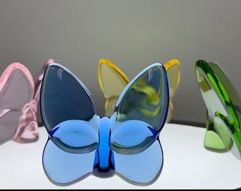 Könnte beinhalten: Eine Sammlung farbenfroher Glas-Schmetterlingsfiguren in Blau-, Rosa-, Gelb- und Grüntönen. Die Schmetterlinge sind auf einer weißen Oberfläche mit einem weichen, verschwommenen Hintergrund ausgestellt.