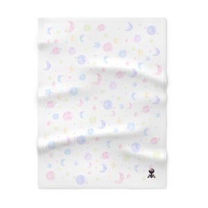 Op de afbeelding: Witte deken met een pastel aquarelpatroon van planeten, sterren en halve manen. Het hemelse ontwerp heeft zachte roze, blauwe, gele en paarse tinten. De deken is opgevouwen en heeft een kleine zwarte afbeelding in de rechter benedenhoek.