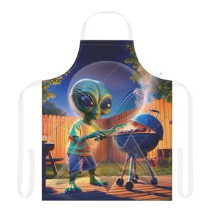 Puede incluir: Delantal con una ilustración caprichosa de un extraterrestre haciendo una barbacoa. El extraterrestre lleva una camiseta amarilla y pantalones cortos azules. El fondo muestra una valla de madera, un cielo estrellado y una barbacoa.