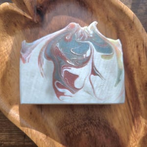 May include: A rectangular bar of handmade soap with a marbled design in shades of white, red, and teal. The soap is resting in a wooden, leaf-shaped bowl. The soap's surface has a swirled pattern.