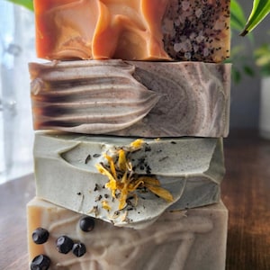 May include: A stack of four handmade soaps with varying textures and colors. The top soap is a caramel color with a swirled design. The second soap is a marbled gray and brown. The third is a pale green with yellow flower petals. The bottom soap is light beige with dark berries.
