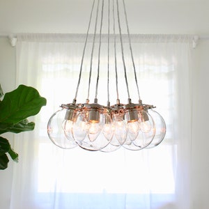 May include: A modern, chrome chandelier with clear glass globe light bulbs. The chandelier is hanging from a white ceiling in a room with a white wall and a white sheer curtain.