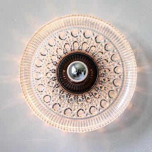 May include: A round, clear glass ceiling light fixture with a decorative pattern. The light has a bronze-colored center and a silver bulb. The fixture is mounted on a white wall.