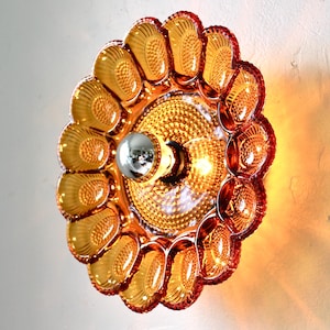 May include: A decorative wall sconce with a flower-like design. The sconce features a central light fixture with a silver bulb, surrounded by amber-colored glass petals. The fixture is mounted on a white wall, casting a warm glow.
