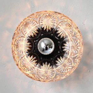 Pode incluir: Uma luminária redonda de vidro transparente com um padrão de estrela. O centro é preto com um globo prateado. A luminária foi projetada para ser montada em uma parede ou teto.