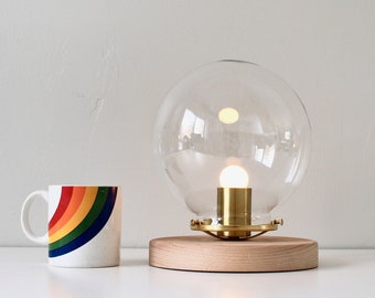May include: A modern table lamp with a clear glass globe shade and a wooden base. The lamp is turned on and the light bulb is visible.