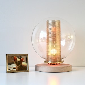 May include: A modern table lamp with a clear glass globe and a brass cylinder with a patterned design. The lamp is on a wooden base and is lit with a warm white light.