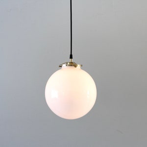 May include: A white globe pendant light with a brass fixture hangs from a black cord against a white wall.