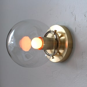 May include: A brass wall sconce with a clear glass globe shade and a light bulb turned on.