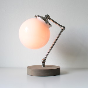 May include: A modern desk lamp with a round, frosted glass shade and a chrome-finished adjustable arm. The lamp sits on a round, gray wooden base. The light is on, illuminating the shade with a soft glow.