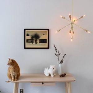 May include: A light wood desk with a drawer, a white ceramic figurine, a vase with dried branches, and a framed black and white print of a snowy landscape. A ginger cat sits on the desk.