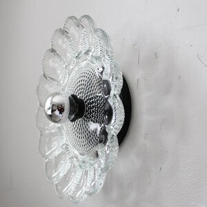 May include: A clear glass wall sconce with a scalloped edge and a textured surface. The sconce has a black metal base and a single light bulb.