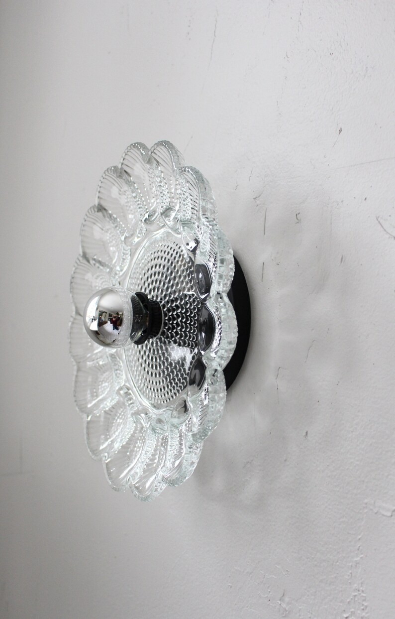 May include: A clear glass wall sconce with a scalloped edge and a textured surface. The sconce has a black metal base and a single light bulb.