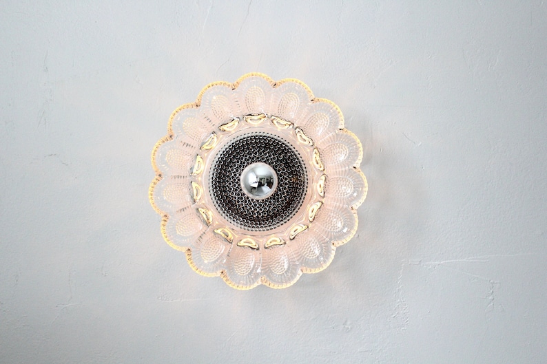 May include: A white and gold ceiling light fixture with a flower-shaped design. The light fixture has a clear glass shade with a decorative pattern.