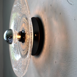 May include: A decorative wall sconce featuring a clear glass plate with an intricate pattern, mounted on a black circular base. The light fixture includes a chrome-tipped bulb, casting a warm glow.