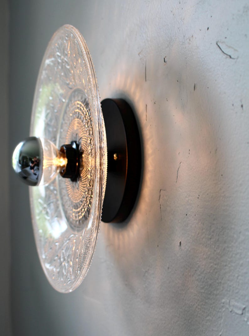 May include: A decorative wall sconce featuring a clear glass plate with an intricate pattern, mounted on a black circular base. The light fixture includes a chrome-tipped bulb, casting a warm glow.