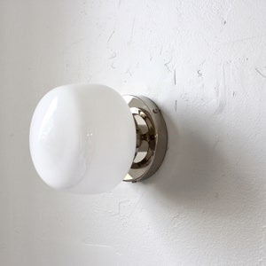 Op de afbeelding: Een witte, gematteerde glazen bolvormige lamp met een chromen metalen voet, gemonteerd op een witte muur.