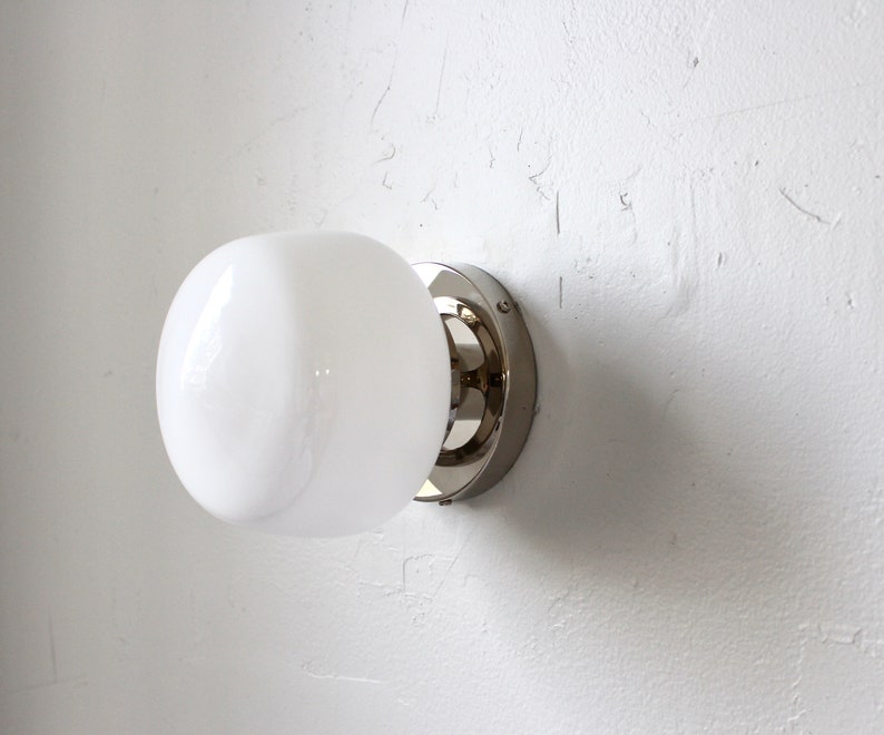 Op de afbeelding: Een witte, gematteerde glazen bolvormige lamp met een chromen metalen voet, gemonteerd op een witte muur.
