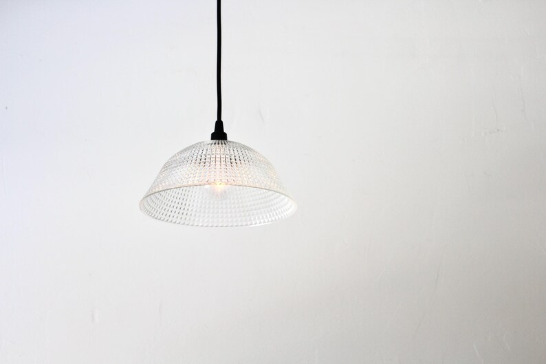 May include: Clear glass pendant light with a textured, dotted pattern shade. The light hangs from a black cord and fixture against a white background. The light bulb is visible inside the shade.