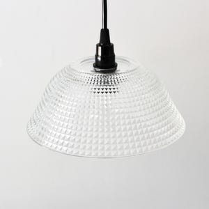 May include: Clear glass pendant light with a textured, geometric pattern on the shade. The light fixture has a black cord and a black fixture base. The shade is a bell shape.