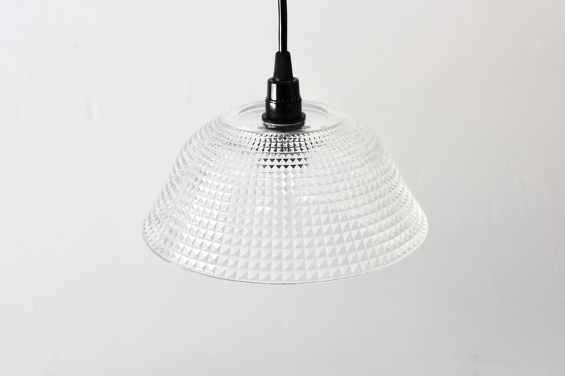 May include: Clear glass pendant light with a textured, geometric pattern on the shade. The light fixture has a black cord and a black fixture base. The shade is a bell shape.