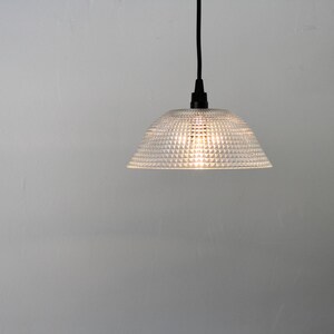 May include: A clear, textured glass pendant light with a dome-shaped shade. The light fixture hangs from a black cord and illuminates a soft glow. The shade has a repeating pattern of small, raised squares.