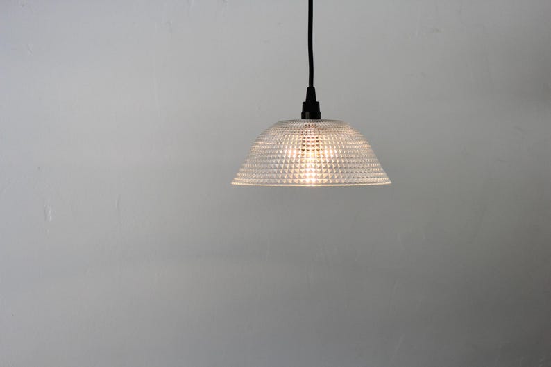 May include: A clear, textured glass pendant light with a dome-shaped shade. The light fixture hangs from a black cord and illuminates a soft glow. The shade has a repeating pattern of small, raised squares.