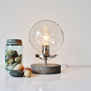 May include: A modern table lamp with a clear glass globe shade and a wooden base. The lamp is lit with a warm white light bulb.