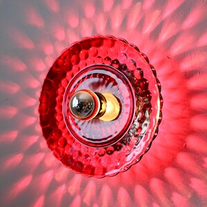 K&ouml;nnte beinhalten: Rote Glas-Wandleuchte mit einem strukturierten, blasenartigen Muster. Die Leuchte hat eine zentrale Gl&uuml;hbirne und wirft ein rotes Lichtmuster an die Wand. Die Leuchte ist ein dekoratives Beleuchtungselement.