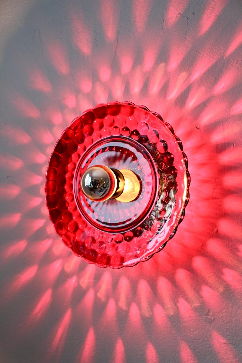 K&ouml;nnte beinhalten: Rote Glas-Wandleuchte mit einem strukturierten, blasenartigen Muster. Die Leuchte hat eine zentrale Gl&uuml;hbirne und wirft ein rotes Lichtmuster an die Wand. Die Leuchte ist ein dekoratives Beleuchtungselement.