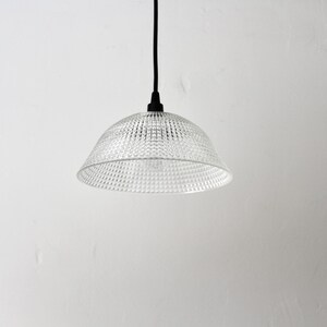 May include: A clear, textured glass pendant light with a bell-shaped shade. The light fixture hangs from a black cord against a white wall. The shade has a patterned surface, and the light bulb is visible.