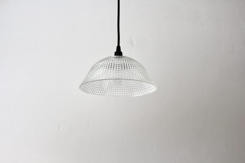 May include: A clear, textured glass pendant light with a bell-shaped shade. The light fixture hangs from a black cord against a white wall. The shade has a patterned surface, and the light bulb is visible.