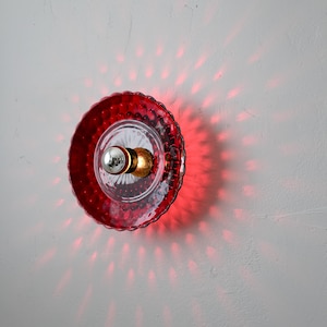 K&ouml;nnte beinhalten: Rote Wandleuchte aus Glas mit strukturiertem, gewelltem Rand. Die Leuchte hat eine klare Mitte mit einer silbernen Gl&uuml;hbirne und einer goldenen Halterung. Rotes Licht strahlt auf die Wand und erzeugt ein Punktmuster.