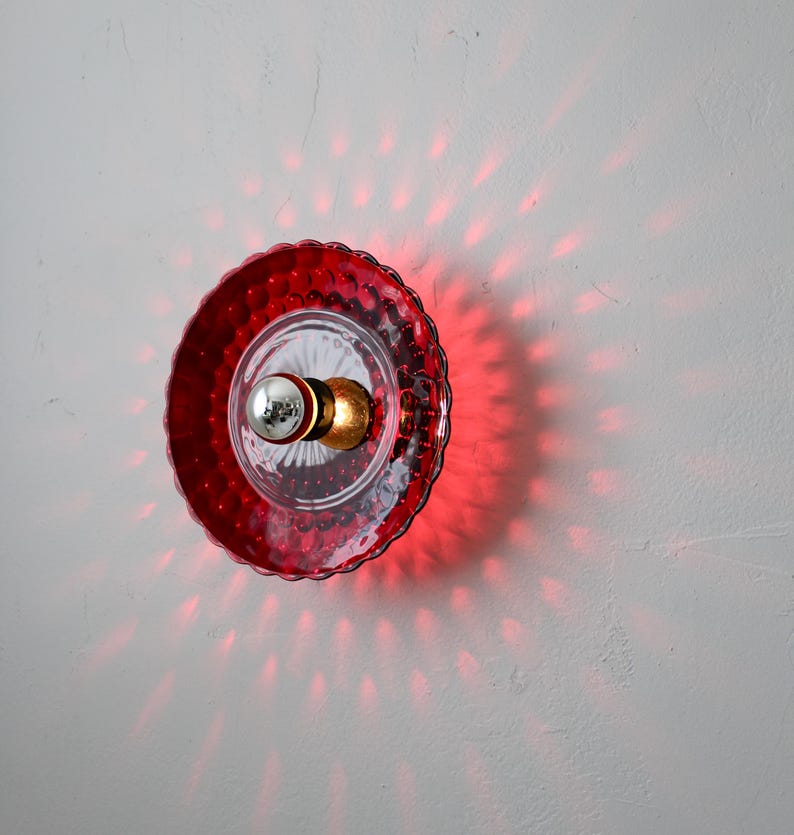K&ouml;nnte beinhalten: Rote Wandleuchte aus Glas mit strukturiertem, gewelltem Rand. Die Leuchte hat eine klare Mitte mit einer silbernen Gl&uuml;hbirne und einer goldenen Halterung. Rotes Licht strahlt auf die Wand und erzeugt ein Punktmuster.
