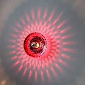 K&ouml;nnte beinhalten: Eine rote, dekorative Wandleuchte mit einem strukturierten, kreisf&ouml;rmigen Design. Die Leuchte strahlt ein hellrotes Licht aus und wirft ein Muster aus Licht und Schatten auf die graue Wand. Die Lichtquelle ist eine kleine, silberfarbene Gl&uuml;hbirne.