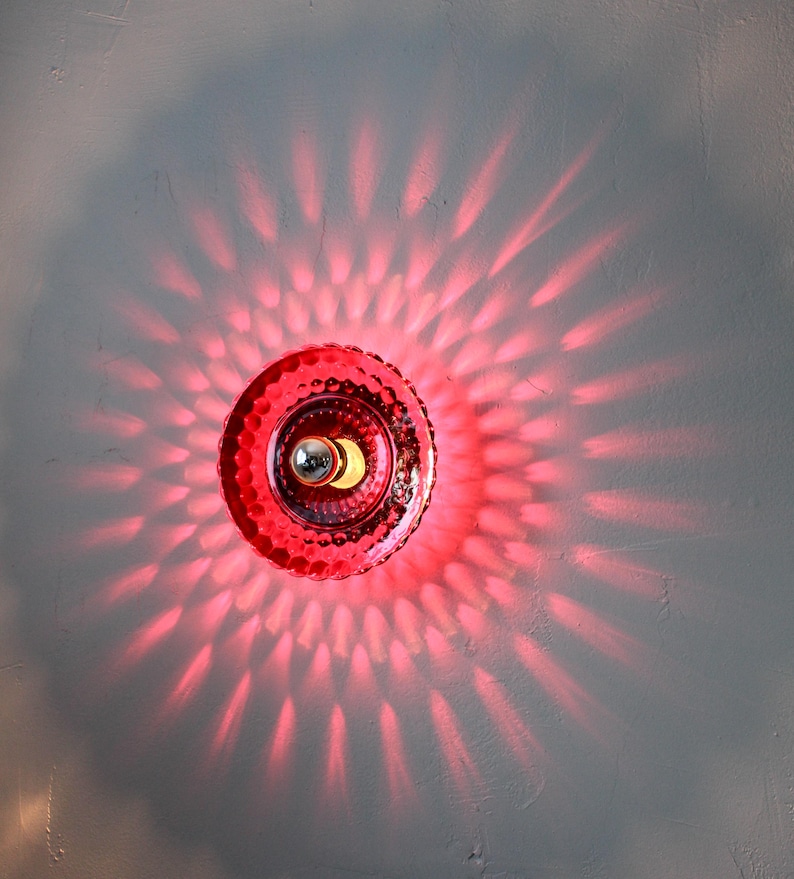 K&ouml;nnte beinhalten: Eine rote, dekorative Wandleuchte mit einem strukturierten, kreisf&ouml;rmigen Design. Die Leuchte strahlt ein hellrotes Licht aus und wirft ein Muster aus Licht und Schatten auf die graue Wand. Die Lichtquelle ist eine kleine, silberfarbene Gl&uuml;hbirne.
