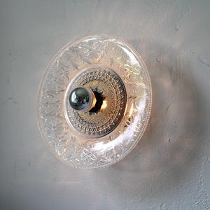 May include: A decorative wall sconce featuring a clear glass plate with floral and scrollwork patterns. The center has a metallic, starburst-like design with a silver bulb. The sconce is mounted on a textured, off-white wall.
