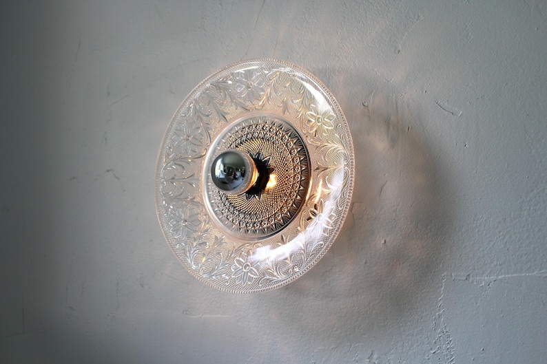 May include: A decorative wall sconce featuring a clear glass plate with floral and scrollwork patterns. The center has a metallic, starburst-like design with a silver bulb. The sconce is mounted on a textured, off-white wall.