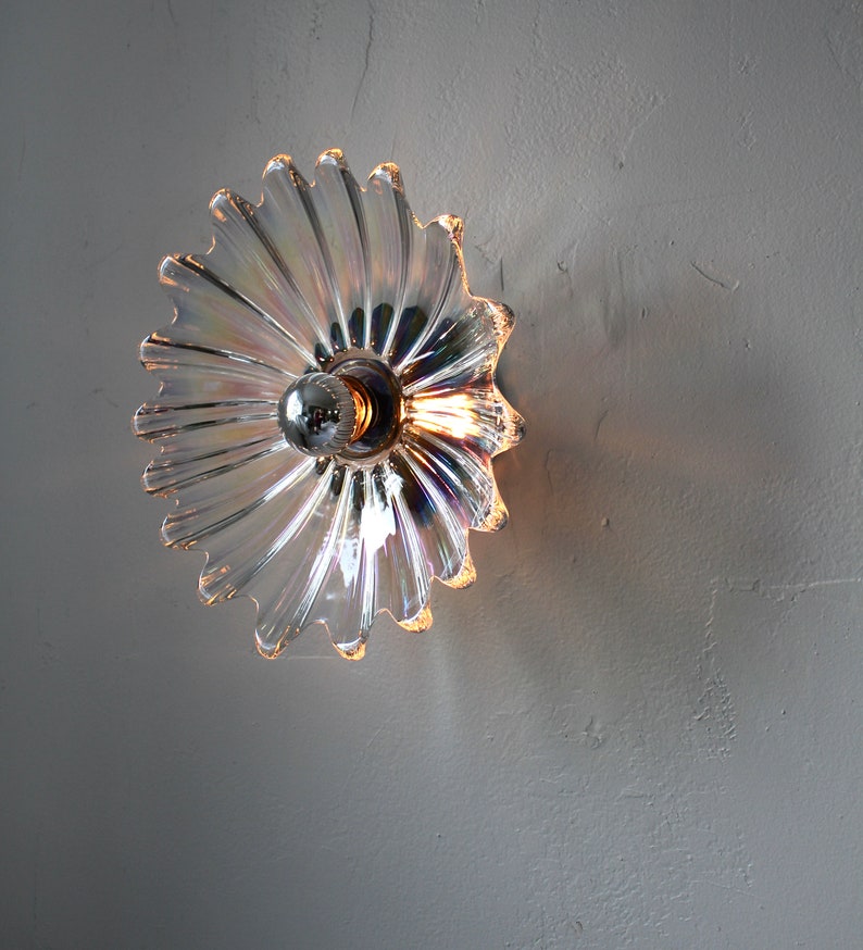 May include: A clear glass wall sconce with a iridescent finish and a silver metal base. The sconce has a flower-like shape with many petals.