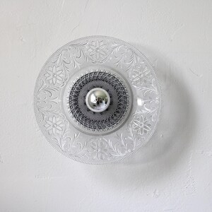 May include: Clear glass ceiling light fixture with a floral pattern around the edge. The center has a silver-colored, round element. The light is mounted on a white wall.