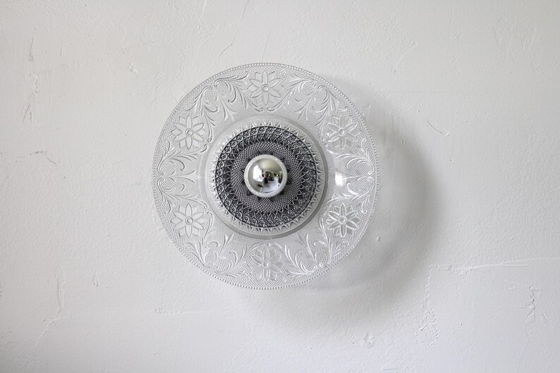 May include: Clear glass ceiling light fixture with a floral pattern around the edge. The center has a silver-colored, round element. The light is mounted on a white wall.