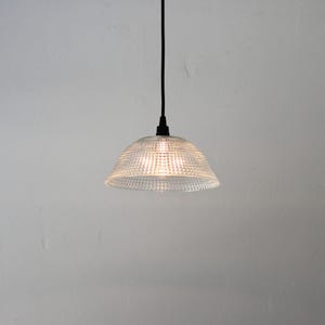 May include: A clear glass pendant light with a textured, dotted pattern. The light fixture hangs from a black cord and is illuminated, casting a warm glow. The light is suspended against a plain white background.