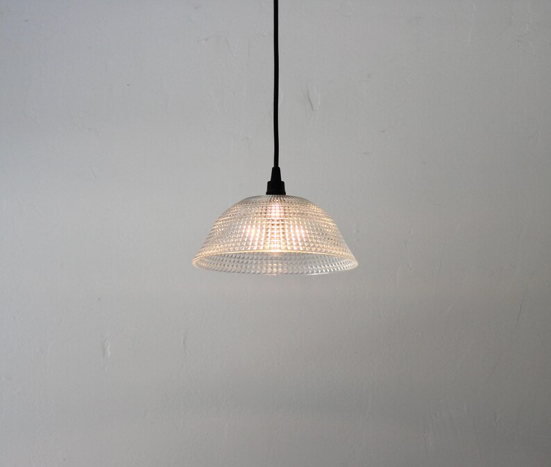 May include: A clear glass pendant light with a textured, dotted pattern. The light fixture hangs from a black cord and is illuminated, casting a warm glow. The light is suspended against a plain white background.